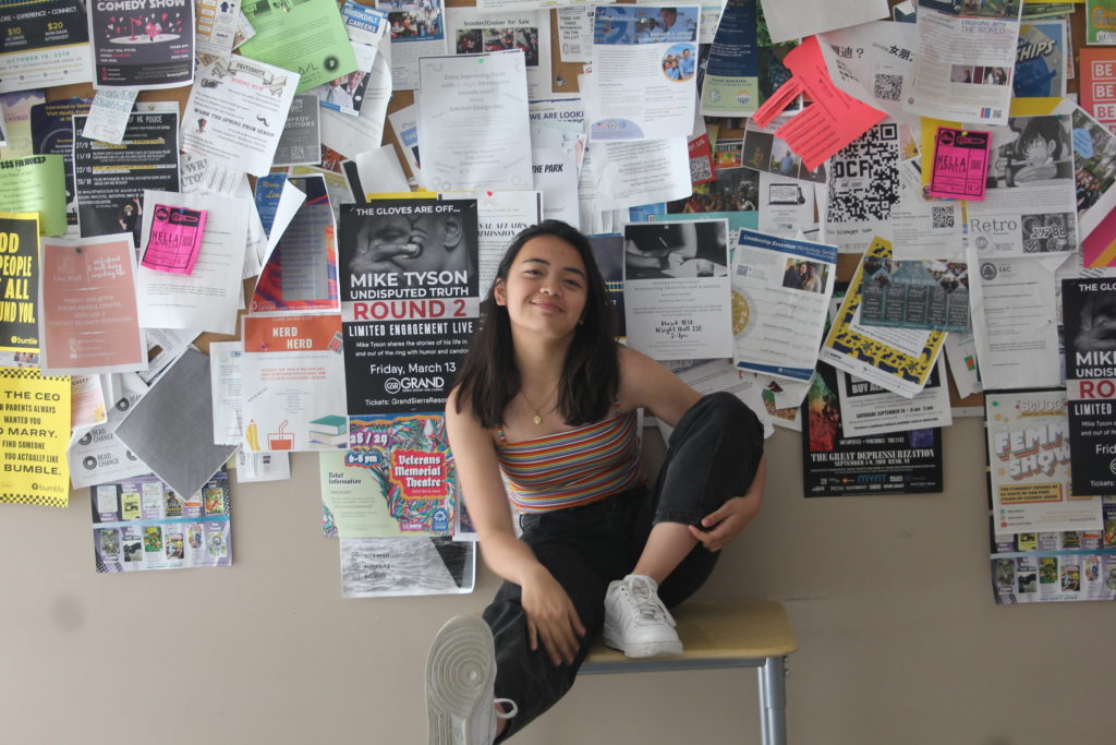 The author is sitting on a stool. Many different papers and advertisements are hung up behind her.