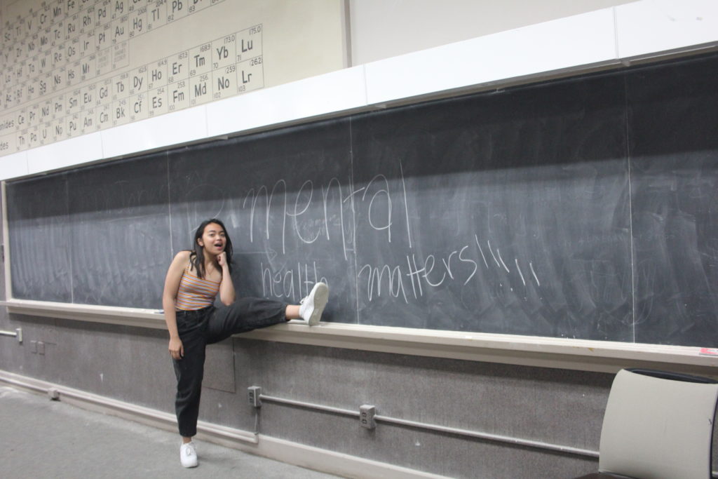 The author standing next to a black board. Mental Health Matters is written on the board in chalk.
